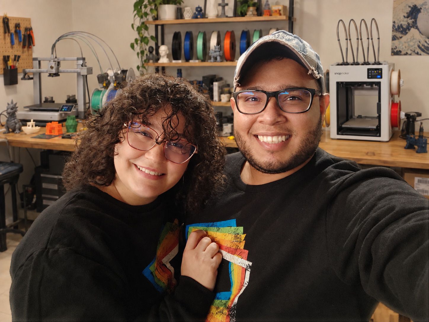Gabriel Salazar y Adriana Adrian sonríen en una selfie dentro de un taller, con dos impresoras 3D y bobinas de filamento detrás.