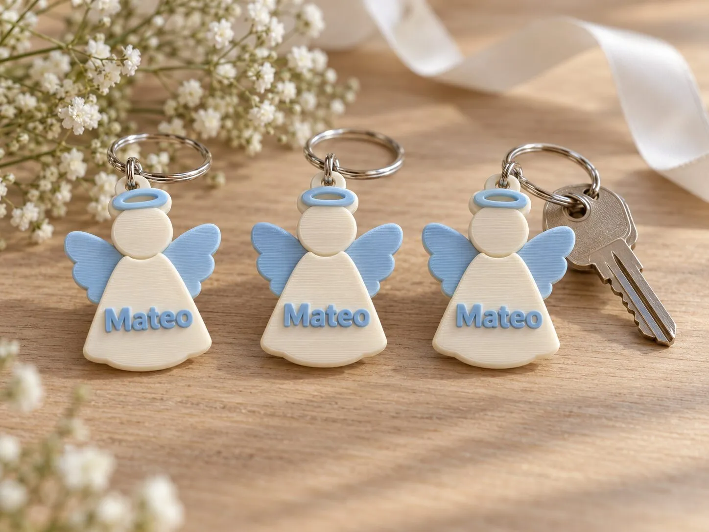Tres llaveros con forma de angelito, de color crema con alas celestes y el nombre Mateo; uno cuelga de unas llaves.