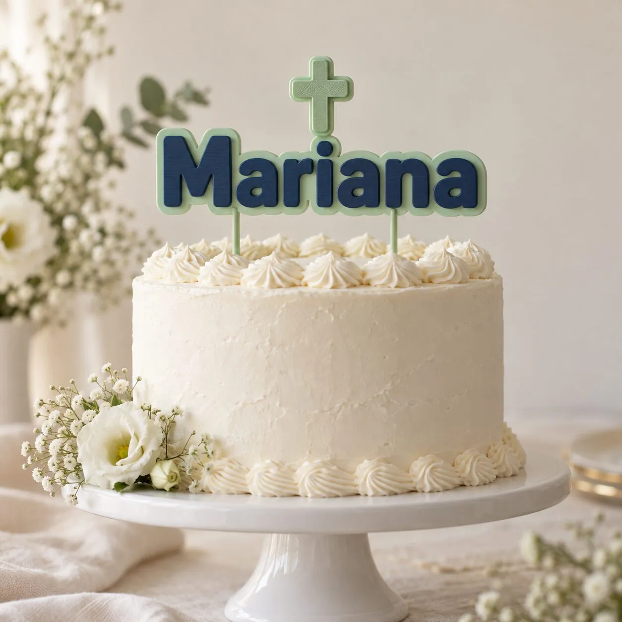 Una torta blanca con un topper que dice «Mariana» en letras azules, con una cruz verde menta encima.