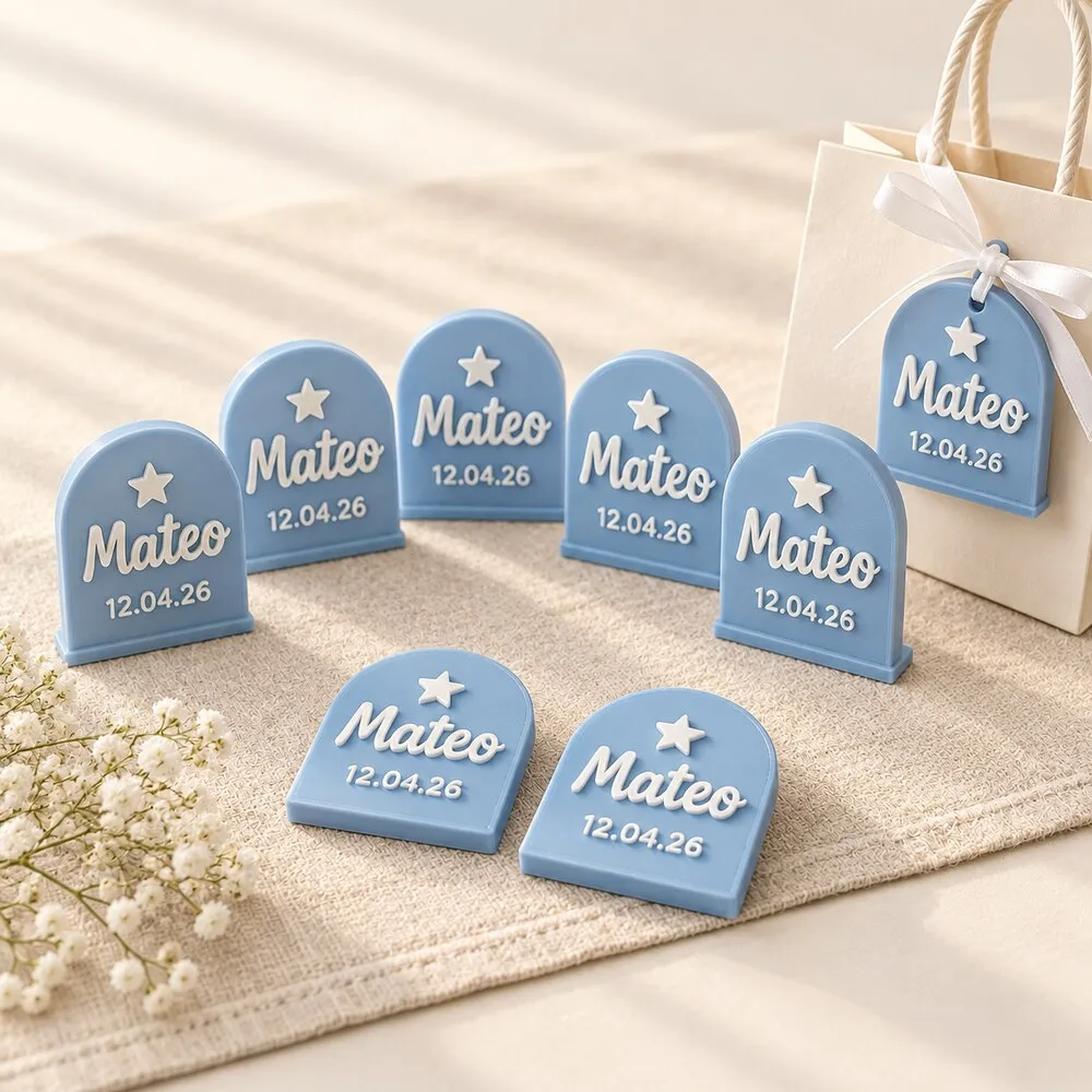 Recuerdos de bautizo: placas celestes en forma de arco con el nombre Mateo y la fecha 12.04.26 en relieve blanco, de pie sobre un camino de mesa de lino, y una más atada con cinta a una bolsita de regalo.